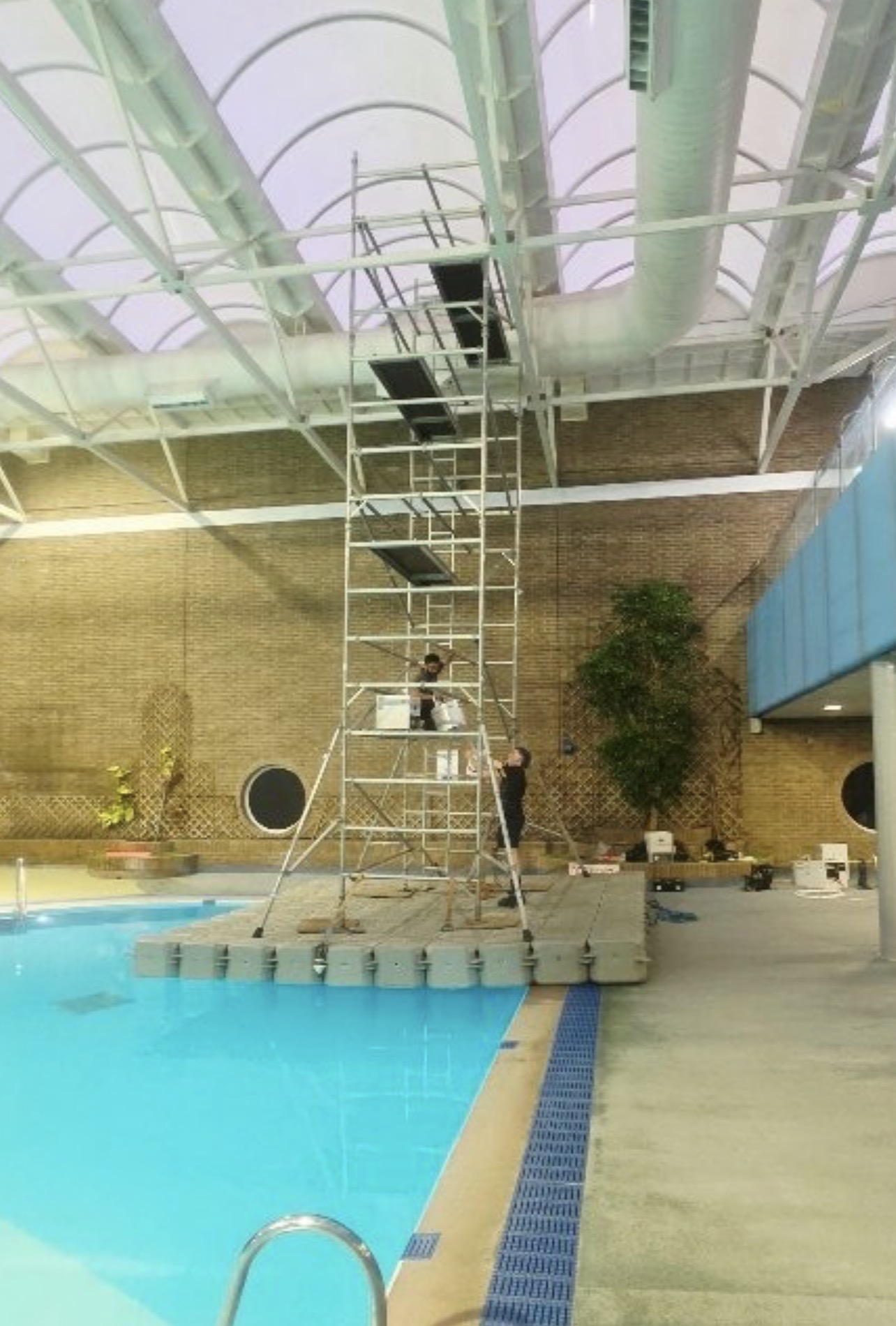 Tall scaffold tower supported by a modular floating pontoon for air circulation fan installation above an indoor swimming pool