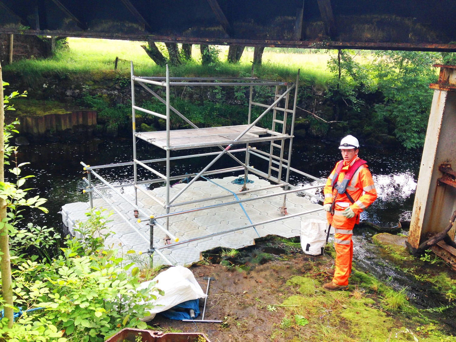 FPS modular inspection pontoon with handrails and a scaffold tower moored at a riverbank beneath a bridge, with an operative in orange hi-vis, hard hat and lifejacket, during the North Lincolnshire bridge inspection programme