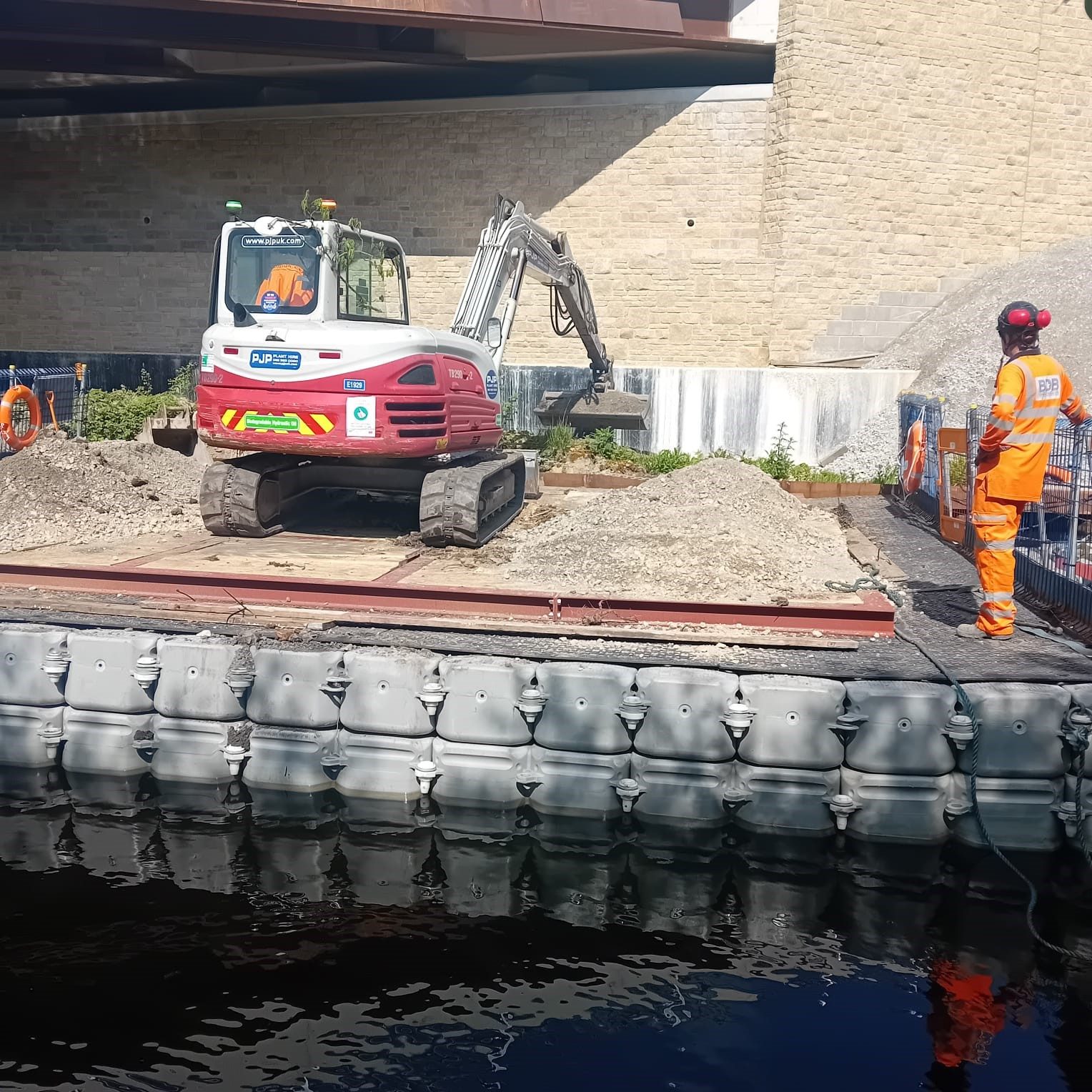 Tracked excavator working on an FPS heavy duty modular pontoon platform with timber decking and edge protection beside a bridge abutment, with an operative in orange hi-vis, during A629 Halifax bridge works